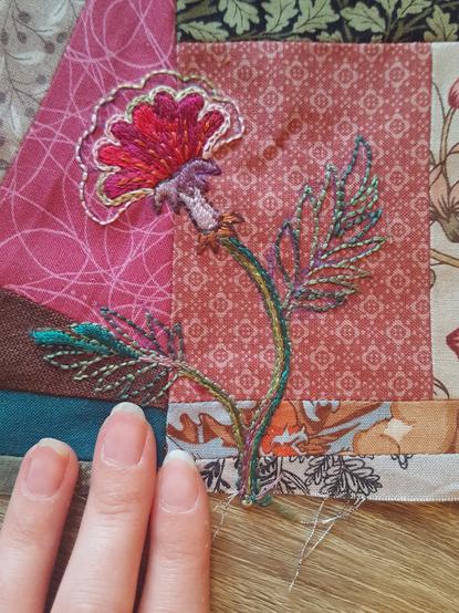 Top down photo of an old fashioned shape flower embroidered on a patchwork block. It's done in vibrant mixed hues, and vaguely has the look of traditional embroidery and motifs on folk dress and 18th century inspired nature prints, but due to the colours and background looks pretty modern also. An outline has been backstitched in variegated embroidery floss, in four different colourways, and this has then been partially filled in with chain stitch, satin stitch, straight stitch and split stitch, giving it a bunch of different textures that nonetheless go nicely together. One leaf of two has a bit of satin stitch fill in it, in a vibrant teal pine green, matching the little patch of fabric right below it. It's not a grand piece of art, but it looks fairly intentional and dynamic all in all! Sini's fingertips in frame, she has shortened her nails.