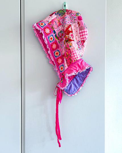 A colorful, patchwork hood with floral and geometric patterns in shades of pink and purple is hanging on a wall. It features ruffled edges and long ties.