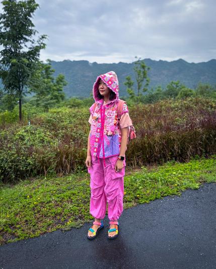 A person stands outdoors in a vibrant pink outfit featuring a hooded vest and wide pants. The background includes lush greenery and distant mountains under a cloudy sky.