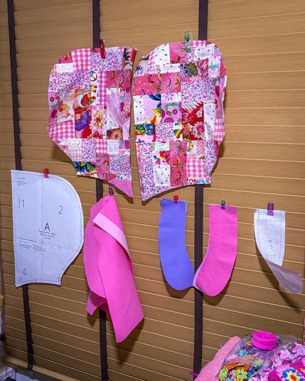A sewing workspace featuring colorful quilted fabric pieces clipped to a wall, along with various sewing patterns and fabric shapes in pink and purple. The background shows wooden blinds, and there are craft materials visible at the bottom.
