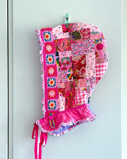 A colorful, patchwork hood in shades of pink hangs against a white wall. It features various floral and patterned squares, decorated with ruffled edges and colorful ties. A clip holds it in place.