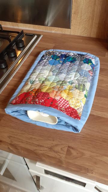 A hot water bottle cover. The front is made of colourful hexagons of different fabrics arranged in rainbow order. The edge is bound with a strip of pale blue fabric. A cream coloured hot water bottle is tucked inside.