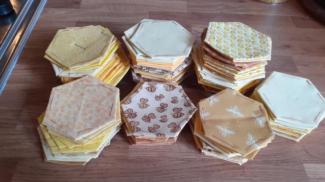 7 stacks of fabric hexagons, each tacked to a hexagon of paper, for  piecing together. The hexagons are made of a selection of fabrics, all with a yellow or golden colour. Some are plain, some have patterns, including some with bees on and some with fabric that has a honeycomb type pattern.