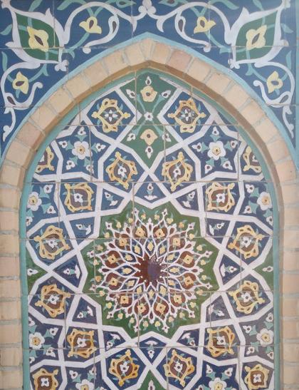 Detail of an exterior wall showing an elaborate tiling. A pointed arch frames a rosette of white lines with green and blue cells filled with yellow floral and green vegetal elements.