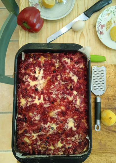 A large tray bake with cheesecake and red berry topping, resting on a table to cool. A lemon grater and half lemon sit on the breadboard to the right, various plates and knives above.