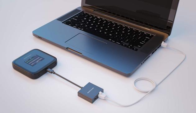 Ein MacBook steht auf einer weißen Oberfläche und ist mit einem Ethernet-Kabel und einem USB-C-auf-Ethernet-Adapter mit dem Netgear Nighthawk M3 Router verbunden. Foto (c) Netgear.