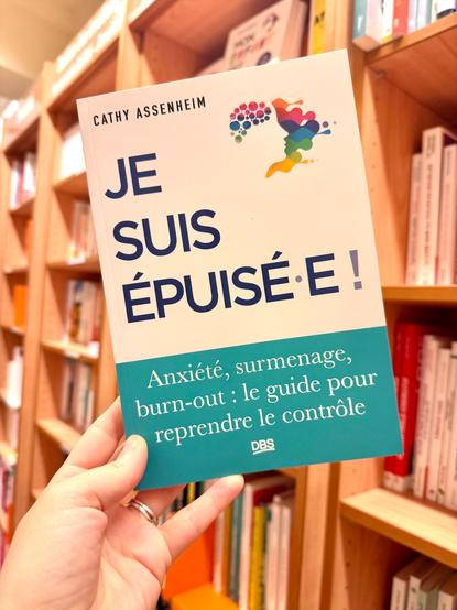 Livre pris en photo devant un mur d’étagères pleines de livre. La couverture est blanche avec un logo de visage de profil dans des taches de couleur.