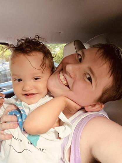 A light-skinned biracial baby with dark curly hair smiles slightly and holds their hand up to the side of their parent's face. The parent, who is white, has their head tilted to the side with a big smile, and has short light brown hair. The background is the inside of a vehicle