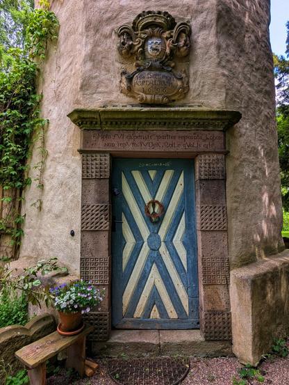 A full-frame, eye-level shot captures the entrance to a round, stone tower, possibly a historical folly or small church, nestled amongst green foliage. The tower is made of rough-hewn, light brown stone. Above a blue and white painted wooden door, an ornate stone relief features a coat of arms and an inscription, partially obscured, but with visible text "1696". Below it, another inscription is carved into the stone lintel of the doorframe. The door itself is painted in a striking geometric pattern of alternating blue and off-white stripes, forming an X-shape within a circle. A small wreath hangs from the centre of the door. To the left of the door, an old wooden bench holds a terracotta pot overflowing with purple and white flowers. Green ivy climbs the tower walls on the left side of the image, while on the right, more foliage and the base of another stone structure are visible. The overall impression is one of rustic charm and historical significance.