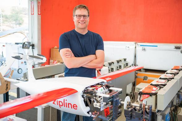 A photo of Keenan standing next to a white and orange Zipline drone. He's wearing a dark blue t-shirt and glasses.