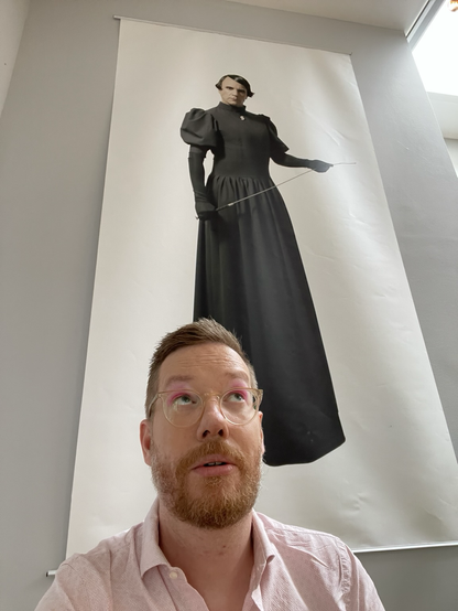 A picture of Redfern gazing up at a large poster of a strict and genderqueer schoolmistress-looking figure. Redfern is wearing a light pink shirt and hot pink eyeshadow like the girlboss they are.