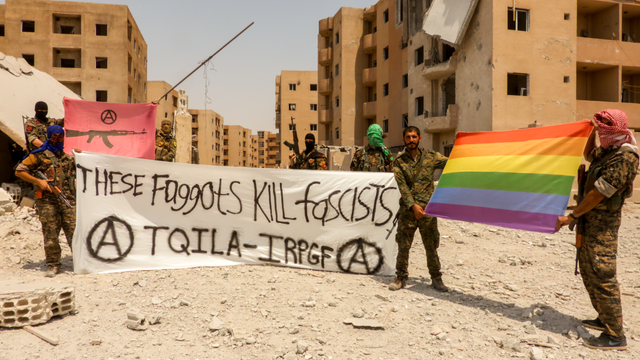 TQILA in Raqqa. Image shows heavily armed TQILA fighters in fatigues, some wearing balaclavas or face coverings, standing in front of shelled out buildings, displaying the Pride Flag, a red flag with an M16 and a circle “A,” and a banner that reads: These Faggots Kill Fascists, with 2 circle “A’s.” By IRPGF, CC BY-SA 4.0, https://commons.wikimedia.org/w/index.php?curid=89781661