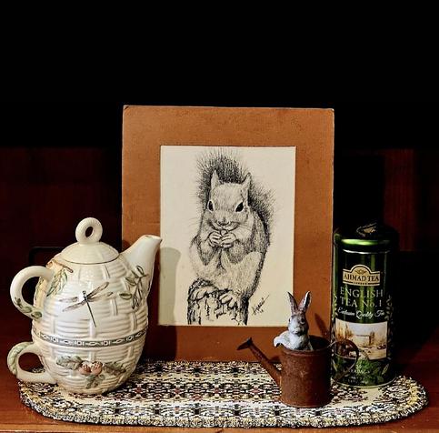 Cozy tea tableau in warm tones centered around a vintage pen and ink drawing of a squirrel eating a nut. The service is a Naturewood tea for one set by Pfaltzgraff featuring pinecones and a dragonfly on pale ceramic etched to look like basketry. Nearby is a rusted miniature metal watering can with a rabbit figurine popping up from within. The tea is English black blend Number 1 from Ahmad. All are set upon a small, vintage tapestry runner.