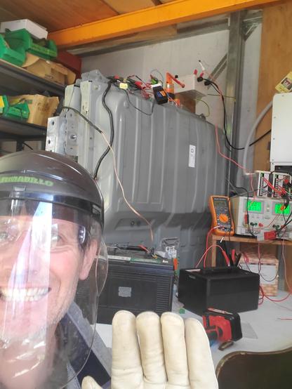 Man in face shield, safety glasses and class 00 HV gloves grins at camera. In background are a variety of power supplies and batteries all connected to a Nissan Leaf battery.