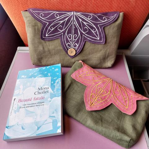 Sur la tablette rose d'un siège de train se trouvent deux pochettes en lin vert, avec des rabats en forme de fleur. La plus grande a un rabat violet avec des broderies d'arabesques argentées, l'autre a un rabat rose avec des broderies dorées. 
Elles se ferment grâce à un bouton en noix de coco. 

Ce sont des pochettes à livre. La plus petite pochette contenait "beauté fatale, les nouveaux visages d'une aliénation féminine" de Mona Chollet.