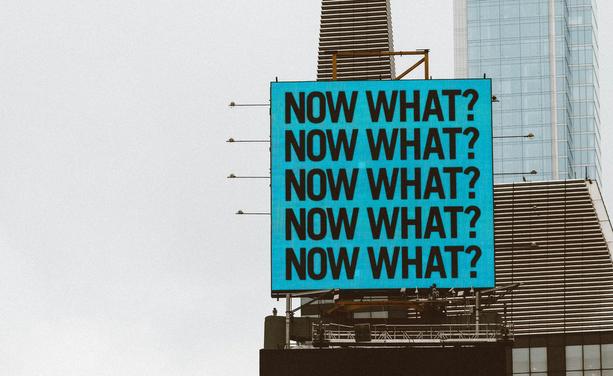 Blue sign on a building with 5 Now What? written
