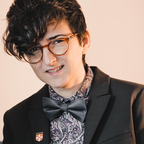 A photo of Didem looking happy, wearing a black a white patterned shirt, a suit jacket and bow tie.