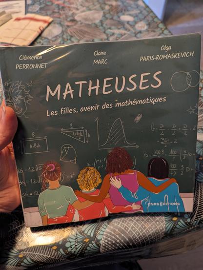 Photo du livre mentionné dans le pouet. La couverture représente de jeunes filles aux couleurs de peau et de cheveux variées, bras dessus bras dessous, face à un tableau d'école recouvert de figures et d'équations mathématiques.