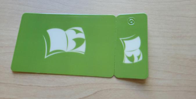 Photo of plastic library card and attached small keychain library card. The designs show an illustration of an opened white book with the letters "LG" displayed in the negative space formed by pages in shadow (at left) and the back of a page with its corners curled (at right). The background is solid yellow-green.