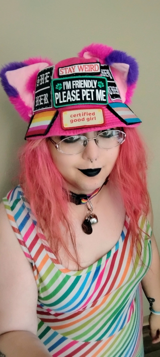 selfie of vanta rainbow black in her iconic hot pink bucket hat with cat ears and patches sewn on and rainbow dress and pink hair and black eyeliner and lipstick. she's smiling a bit. also rainbow collar with a bell! and pink hair