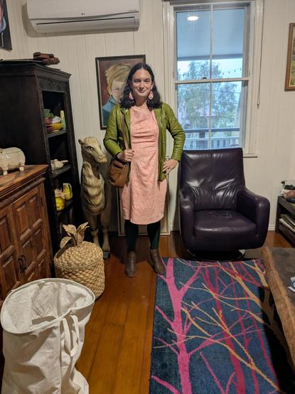 Photo of me wearing the pink dress outfit from the flat lay. I'm standing in a cozy room with wooden floors, a colorful tree-patterned rug, a medium-sized woven basket shaped like a pineapple, and a camel sculpture behind me. A purple leather armchair sits nearby, and rain is visible through the window.