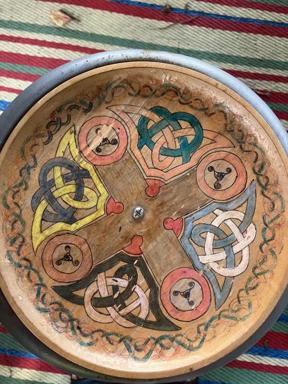 A wooden plate with a quartered design pattern with Celtic knots and triskeles in coloured pencil. A striped mat is seen under it