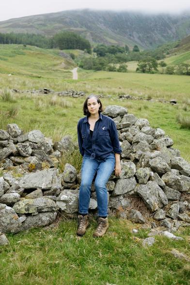 A woman in a navy work shirt and jeans sits on a partially built drystone wall in a green Scottish glen. Behind her, rolling hills and a mist-covered mountain rise in the distance, with a winding path cutting through the landscape. The scene is lush and overcast, evoking a quiet, grounded atmosphere.