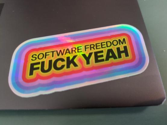 A shiny-backed sticker, generally in the shape of a rounded-corner slanted parallelogram. In the center, black words "SOFTWARE FREEDOM FUCK YEAH" surrounded by concentric outlines of yellow, orange, red, pink, purple, and blue.