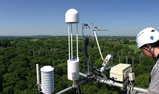 A person in a white safety helmet working at a set of scientific measuring instruments on a platform elevated above a forest in a flat landscape. There are rings of other tall metal structures placed in the forest in the background.