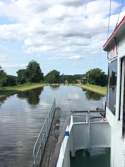 View from the boat deck showing the canal's lock system ahead. Multiple wooden lock gates are visible across the waterway, with green grass areas between them. Trees line both sides of the canal, and there's a small building with a red roof visible in the distance.