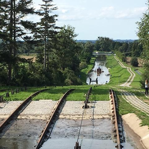 Elevated view looking down at railway tracks running parallel to the canal. The tracks extend into the distance where a boat can be seen in the water. Two people are standing on a concrete platform beside the tracks. Green fields and forests stretch to the horizon under a cloudy sky.