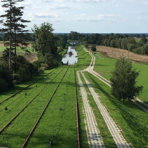 Similar elevated view of the railway system used to transport boats overland. The parallel tracks run straight through green countryside toward a body of water in the distance. Lush forests and agricultural fields are visible on both sides, creating a picturesque landscape typical of the Warmian-Masurian region.