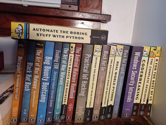 A shelf filled with No Starch Press books on hacking, programming, and computer science. From left to right, the visible spines are:

Metasploit: The Penetration Tester’s Guide (2nd Edition)

Black Hat Bash

The Art of Mac Malware Vol. 2

Bug Bounty Bootcamp

Evading EDR: Defeating Endpoint Detection Systems

The Art of Mac Malware

Linux Basics for Hackers

The Android Malware Handbook

Practical IoT Hacking

Beyond the Basic Stuff with Python

Black Hat Go

The Recursive Book of Recursion

Real World Python

Black Hat GraphQL

Windows Security Internals

Codebreaking

The Complete Developer

R for the Rest of Us

Resting horizontally on top of the books is Automate the Boring Stuff with Python (3rd Edition), its spine facing outward. Most books have the distinctive No Starch yellow-and-black design or mascot illustrations on the spine. The background includes a white wall, a wooden window frame, and a partially drawn white blind.
