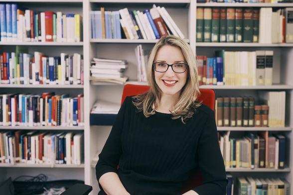 Alena Buys, a mid-aged woman with blond hair and glasses, sitting smiling in front of a book cabinet. She’s wearing a black dress.