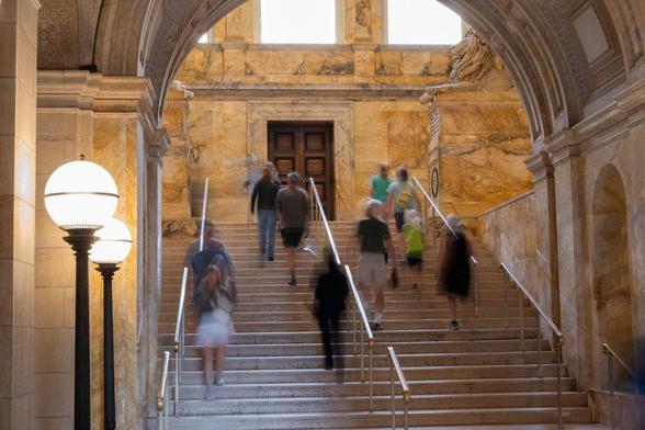 A long exposure photograph inside the Boston Public Library shows people ascending and descending the grand marble staircase. The soft blur of motion gives a sense of timeless activity beneath high arched ceilings and warm stone walls. A stone lion sculpture watches from above.
