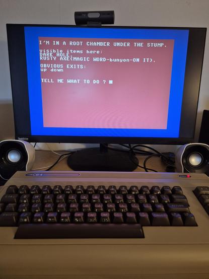 A theC64 computer is set up on a desk in front of a monitor displaying a classic text adventure game. The screen shows a red and blue interface with the text: "I'M IN A ROOT CHAMBER UNDER THE STUMP." Visible items include a "DARK HOLE" and a "RUSTY AXE (MAGIC WORD-bunyon-ON IT)." The game prompts the player with "TELL ME WHAT TO DO?" Two desktop speakers and a webcam are also visible, and the setting suggests a retro gaming or hobbyist environment.