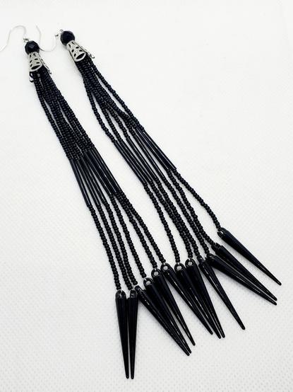 a pair of long, dangling, all-black beaded earrings made by kiamichi. at the top of each earring is a large geodesic clear black bead followed by a silver metal patterned cone opening up to multiple long beaded threads. the threads start with several small shiny black seed beads. in the middle is a long thin shiny black bead with similar thickness to the seed beads followed by more of the same shiny black seed beads. at the end of each thread are long shiny black conical spikes