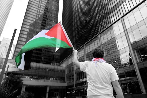 A man with his back turned to the camera, facing up towards a tall building with a Palestine flag in his left hand and a keffiyeh around his neck. The photo is stylized black and white with the flag and keffiyeh in color.
