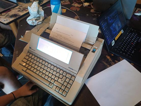 Eine elektrische Schreibmaschine mit Display steht auf einem Tisch, rechts daneben liegt ein Laptop.