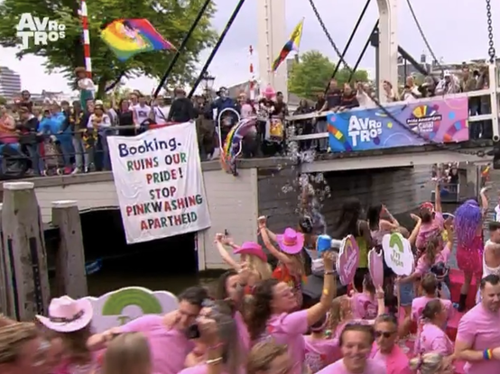 Screenshot NPO 2 AVROTROS live tv uitzending Ansterdam Pride botenparade: Spandoek aan oude brug met de tekst: “Booking. RUINS OUR  PRIDE! STOP PINKWASHING APARTHEID”. Op de voorgrond boot met daarop vooral roze uitgedoste dansende mensen.