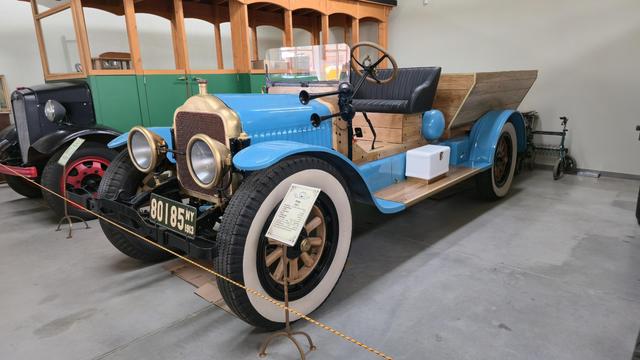 White, a truck by The White Motor Company (US), made in 1913, with white-striped tyres on wooden rims, light-blue panels, wooden constructions around seat and trunk, massive headlights and grille in gold color