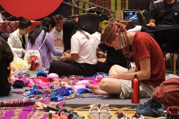 Photo of a maker technology and craft workshop on Peruvian weaving. People are sitting on the floor strongly concentrated on weaving work. In focus is a young white man sitting cross-legged on the floor looking down at their work, which is not visible. Near him are people sitting in a circle, also on the floor, and looking down at their work. There are a lot of colourful Peruvian textiles on the floor, balls of yarn, and shoes. The young man has a woven band wrapped around his head like a headband and a water bottle beside him. Not visible is the information that this is the Sheraton ballroom in Shenzhen where the workshops are taking place. Also not very visible are the small looms made using digital fabrication (laser cutters) which the participants are using to weave their pieces. FAB12, international fab lab conference and meeting, Shenzhen, China, 10 August 2016. Photo Cindy Kohtala