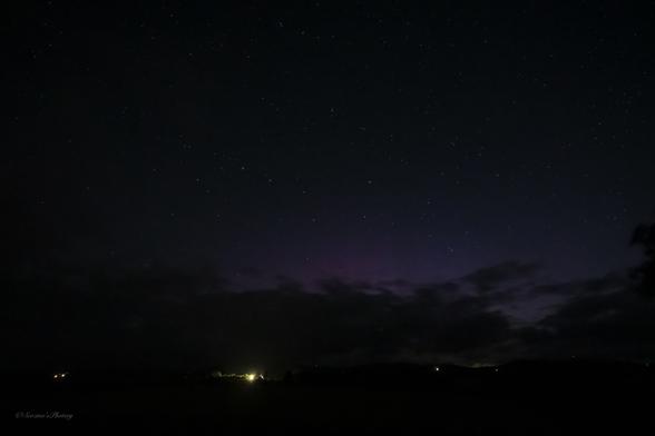 A night sky image with a low bank of cloud over the low dark horizon. Above are stars dotted against a dark sky with pale purple-ish rays showing to the centre. A few house lights show on the horizon