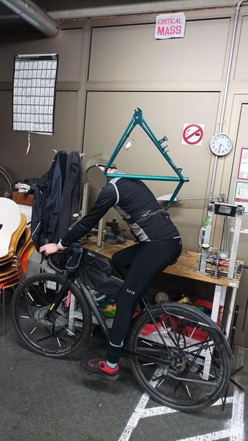 Ein Mann auf einem Fahrrad. Mittels eines Fahrradschlauchs ist ein Fahrradrahmen an dem Mann befestigt, der Rahmen liegt außerdem auf der Schulter des Mannes auf.