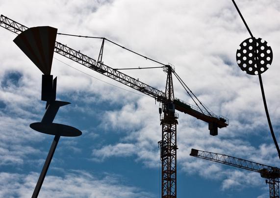 Two thin, tall poles, to the right and left of the photo (part of an art object), were photographed from below and stand out black against the light of the blue and white sky. On the left pole are stacked surfaces, ending in a triangular plane. Against the right pole, halfway up, is a circular surface with large holes in it. In the center is a large crane (also blackened by the backlight). The horizontal section extends diagonally to the right and top left. At the far bottom right is a second, smaller crane in line with the other crane.