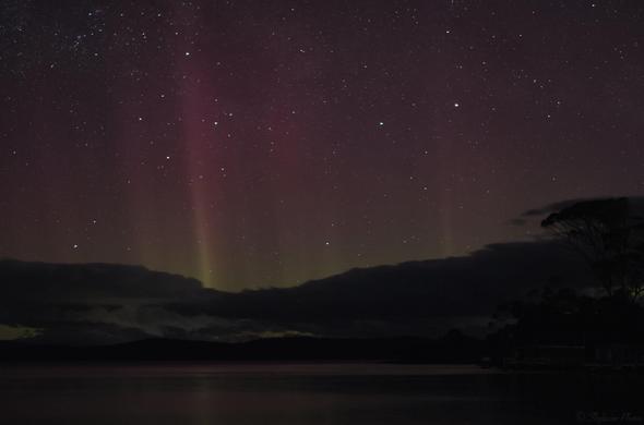 An aurora in the night sky, clouds silhouetted in the distance, faint reflections in the watery foreground.  The colours are faint yellow green near the horizon, turning pale red towards top of the frame.  A bright streak of higher intensity colour beams upwards at mid left, more subtle 'columns' further right.  "The Pointers" (mid upper right) and Southern Cross (mid left) constellations are clearly visible.