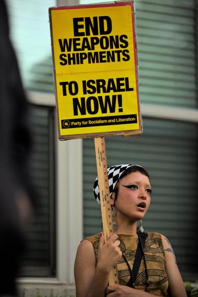 An AFAB person is standing in front of an apartment building, holding a yellow placard that says "end weapons shipments to Israel now!" on the in large letters, underscored by "Party for Socialism and Liberation".

They are outside of Shell Energy Stadium protesting the Kamala Harris rally in the lead-up to the 2024 US presidential elections in opposition to the Democratic Party support of Israel.