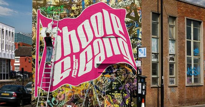 A mural of the fuchsia Phoole and the Gang flag logo covers the entire side of a two-story brick building in a city, painted over many graffiti tags. A painter stands on a ladder completing the painting of our flag onto the wall.