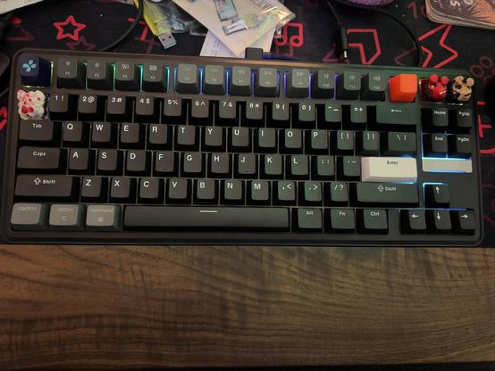 A mechanical keyboard sits atop a richly grained wooden desk, its black frame surrounded by a mix of vibrant artisan keycaps, including red, white, orange, and animal-shaped ones. The backlighting softly highlights some keys in blue and green, illuminating the clean layout. Colorful packaging, USB accessories, and star patterns peek out from the corner of a cluttered background, contrasting with the keyboard's modern design.