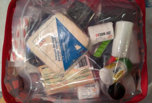 A close view of a soon-to-be first aid kit in a plastic bag inside a red fabric bag. There are rolls of gauze, a triangular bandage, a green nail clipper, and more.
